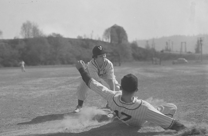 Softball player Margaret Dobson playing for the Vanport College baseball team. She leans downward towards a sliding player, most likely to tag him out. Another player can be seen in the distance, standing on the dirt field. Dobson was inducted into the Oregon Sports Hall of Fame in 1984