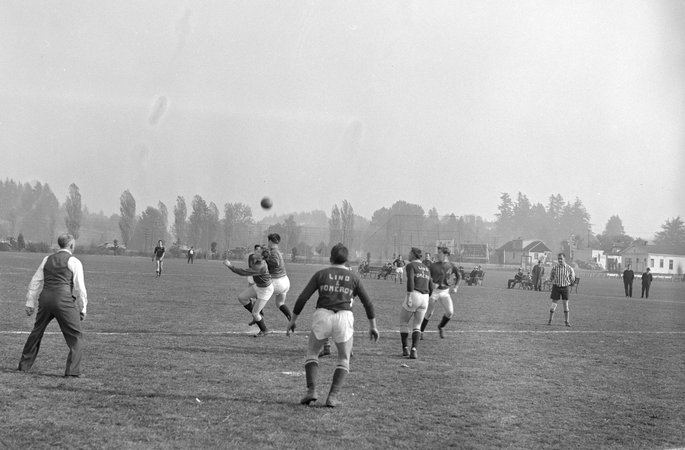 Players for the Albina Engine & Machine Works soccer team, the Subchasers, on a field during a game against a team for Lind & Pomeroy. The ball can be seen in the air, with blurry players underneath